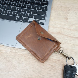 May include: A brown leather card holder with a snap closure and a key ring attached. The card holder is lying on a wooden surface next to a laptop.