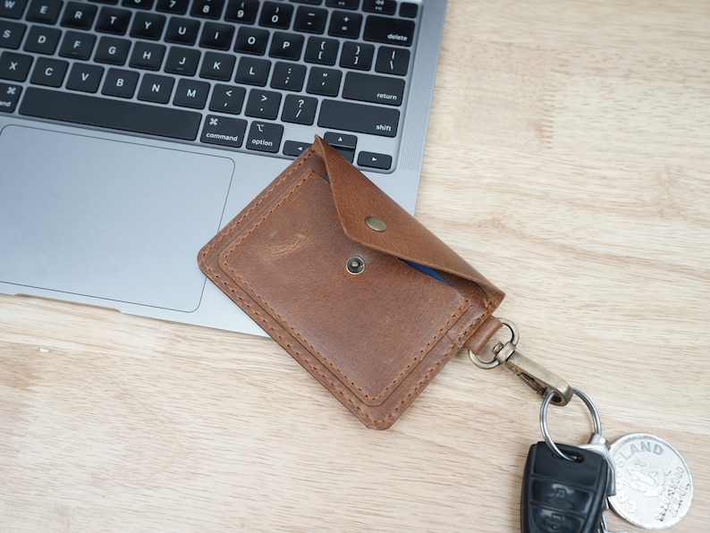 May include: A brown leather card holder with a snap closure and a key ring attached. The card holder is lying on a wooden surface next to a laptop.