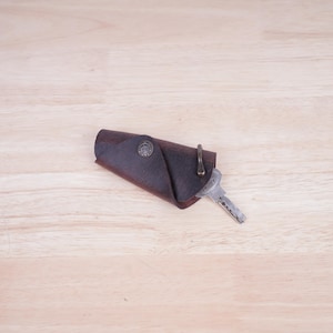 May include: A brown leather key holder with a metal ring and a key attached. The key holder is folded over and has a decorative design on the front.