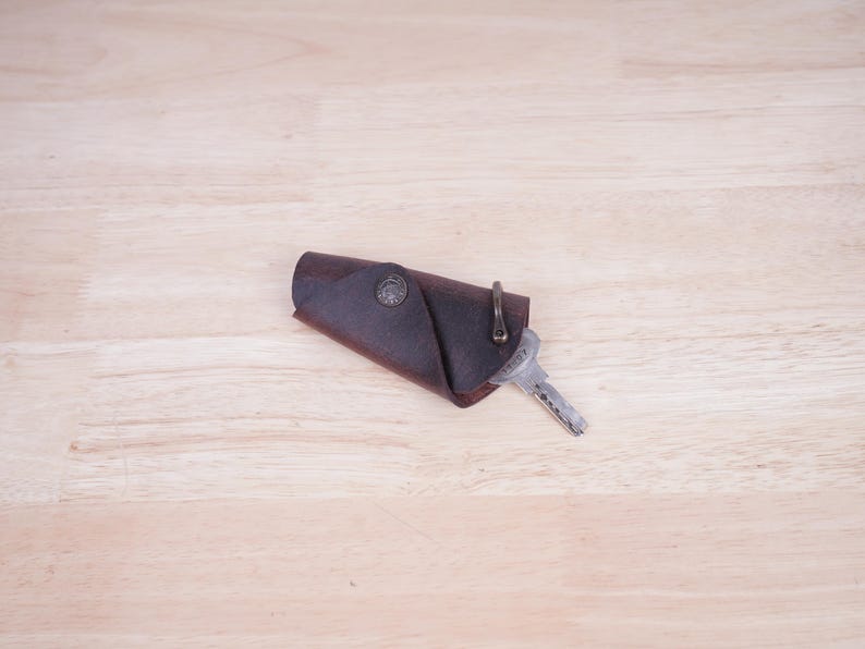 May include: A brown leather key holder with a metal ring and a key attached. The key holder is folded over and has a decorative design on the front.