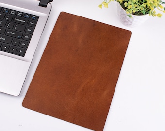 May include: A rectangular brown leather desk mat sits on a white surface next to a laptop and a small potted plant. The mat has a textured surface and appears to be made of genuine leather. The laptop is silver and black.