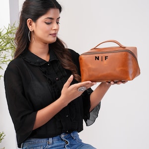 May include: A brown leather travel bag with a top handle and a zip closure. The bag has the initials "N|F" printed on the front. The bag is rectangular and appears to be empty.