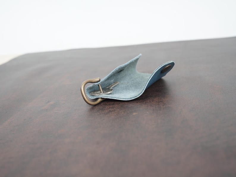May include: A light blue leather key holder with a brass hook and a key inside. The holder is open and the key is visible.