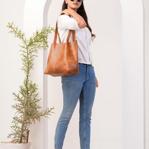 May include: A woman wearing a white button-down shirt, blue jeans, and brown leather tote bag. She is wearing sunglasses and clear heeled sandals.