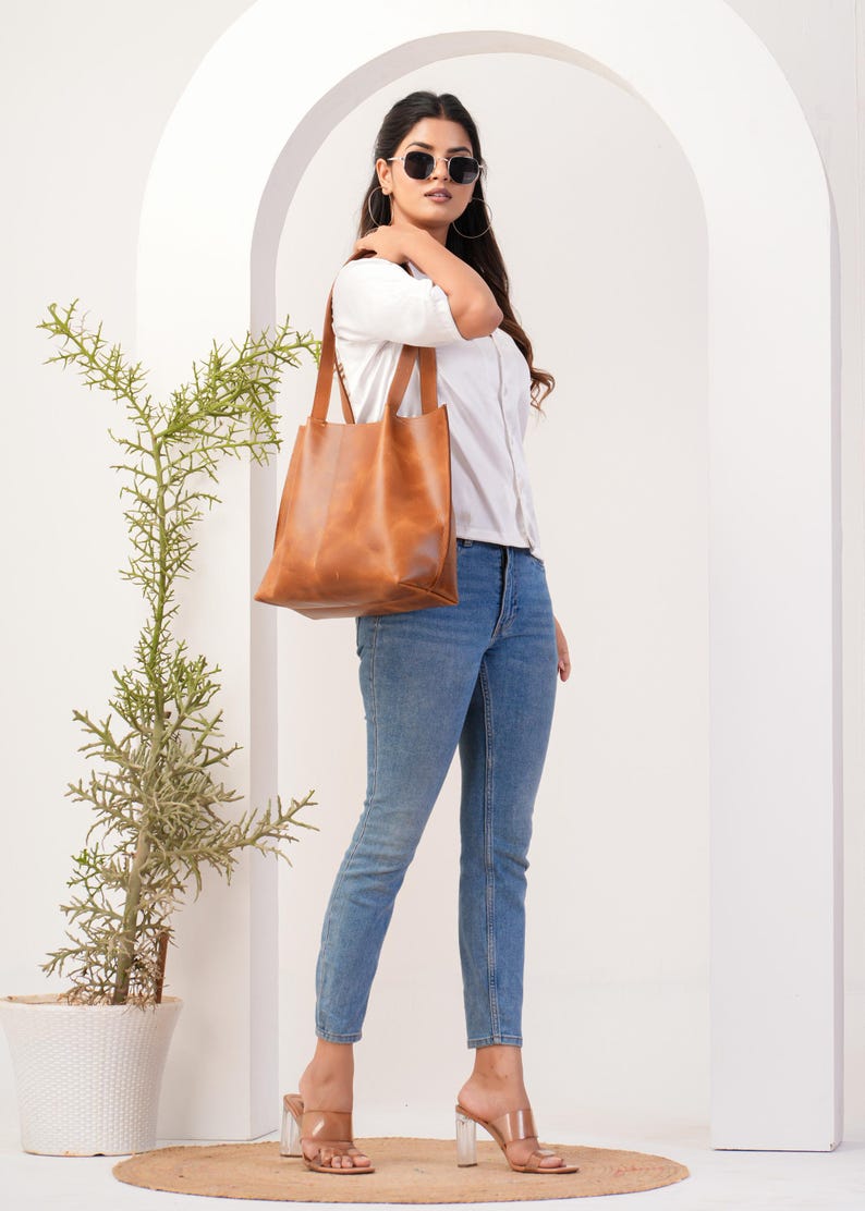 May include: A woman wearing a white button-down shirt, blue jeans, and brown leather tote bag. She is wearing sunglasses and clear heeled sandals.