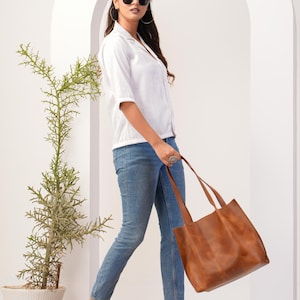 May include: A woman wearing a white button-down shirt, blue jeans, and clear heeled sandals is holding a brown leather tote bag.