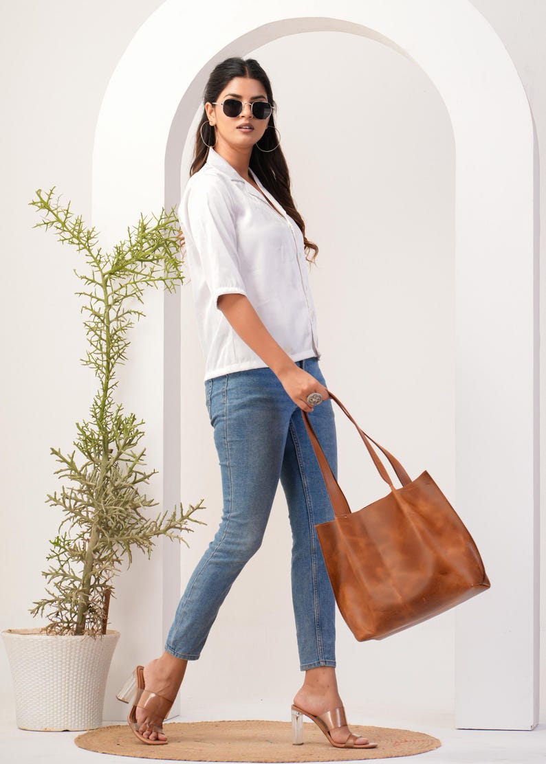 May include: A woman wearing a white button-down shirt, blue jeans, and clear heeled sandals is holding a brown leather tote bag.