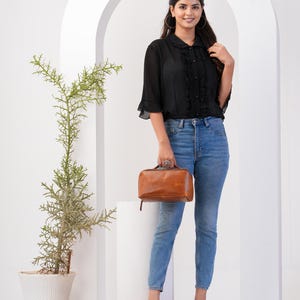 May include: A woman wearing a black ruffled blouse with a brown leather handbag. She is wearing blue jeans and brown heeled sandals.