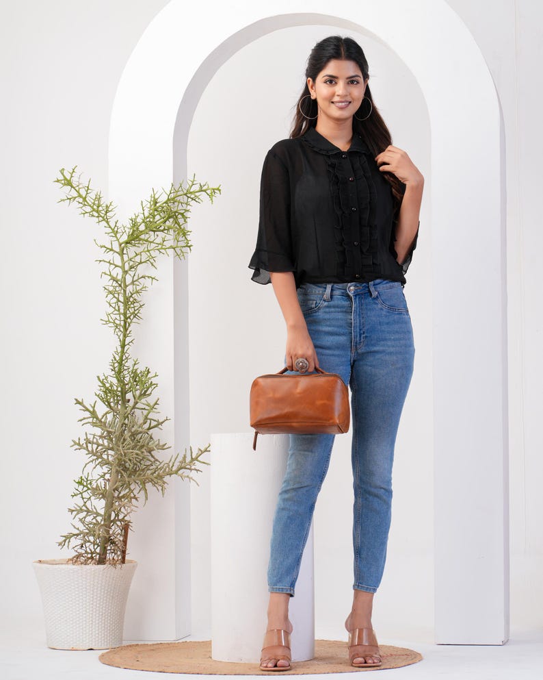 May include: A woman wearing a black ruffled blouse with a brown leather handbag. She is wearing blue jeans and brown heeled sandals.