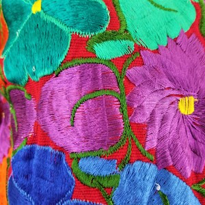 May include: Close-up of colorful embroidered flowers on a red fabric background. The flowers are in shades of purple, blue, green, and yellow.
