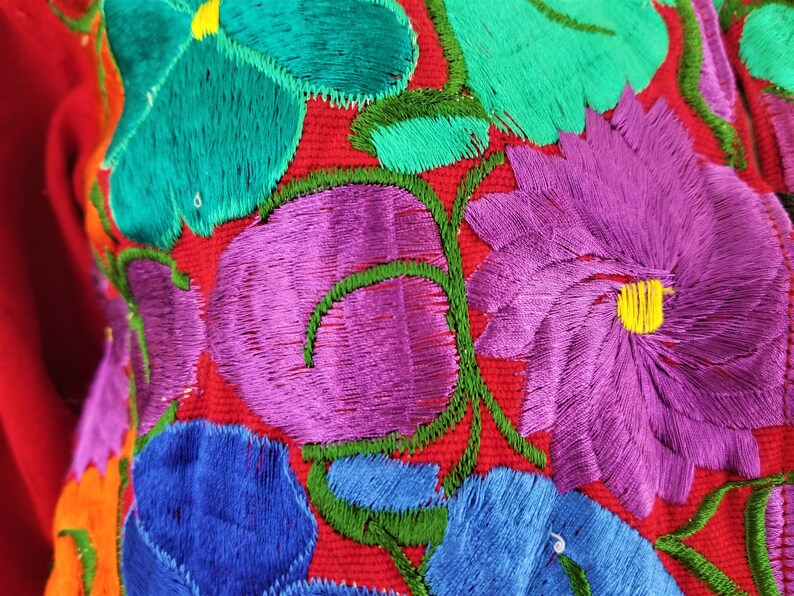 May include: Close-up of colorful embroidered flowers on a red fabric background. The flowers are in shades of purple, blue, green, and yellow.