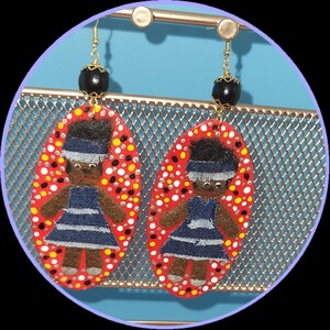 May include: A pair of handmade earrings featuring a red oval design with a pattern of white, yellow, and black dots. Each earring has an embroidered figure wearing a blue dress and a black bead at the top, suspended from gold-colored hooks.