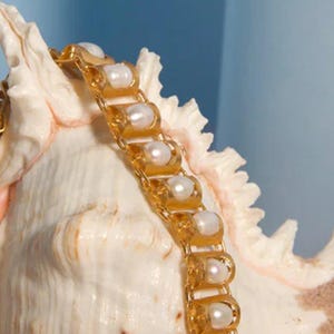 Puede incluir: Una pulsera de metal dorado con una serie de eslabones arqueados repetitivos, cada uno con una perla blanca redonda. La pulsera est&aacute; colocada sobre una concha marina de color crema con textura, un borde puntiagudo y estriado.