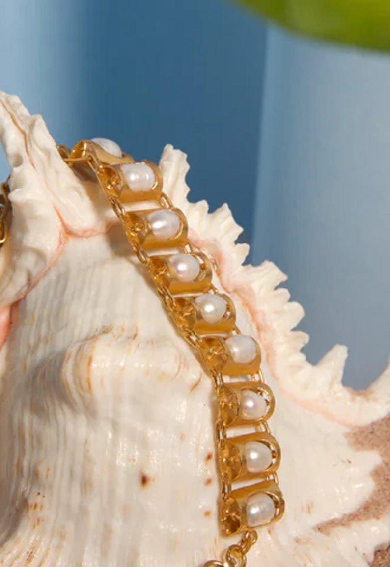 Puede incluir: Una pulsera de metal dorado con una serie de eslabones arqueados repetitivos, cada uno con una perla blanca redonda. La pulsera est&aacute; colocada sobre una concha marina de color crema con textura, un borde puntiagudo y estriado.