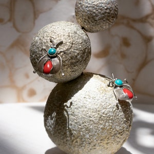 May include: A sculpture composed of three textured, spherical, stone-like forms stacked vertically. Two silver beetle-shaped pins are attached to the spheres, each featuring a small round turquoise stone and a larger red oval stone.