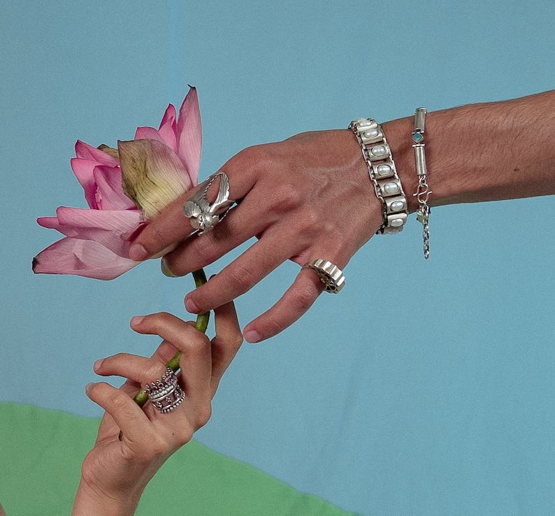 Puede incluir: Un anillo de plata con forma de p&aacute;jaro, una pulsera de plata con piedras blancas ovaladas, una pulsera de cadena fina de plata con una peque&ntilde;a piedra azul, un anillo de plata con borde festoneado y un anillo de plata con piedras rosas sostienen una flor de loto rosa.