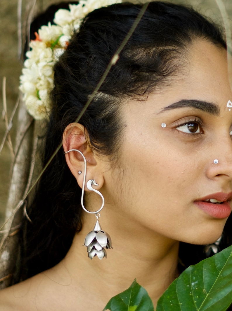 May include: A woman with long black hair wears a silver earring with a swan design and a dangling flower-shaped charm. The earring is worn in her upper ear.
