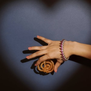 Könnte beinhalten: Eine Hand hält ein süßes, frittiertes Gebäck namens Jalebi, mit einem silbernen Armband mit rosafarbenen Edelsteinen am Handgelenk.