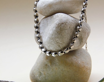 Puede incluir: Un collar de cadena plateada que presenta una serie de pequeñas cuentas metálicas redondeadas unidas entre sí, drapeado sobre una pila de tres piedras lisas de color claro, con un cierre de gancho visible en la parte superior.