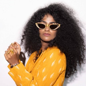 Puede incluir: Una mujer con el pelo negro rizado lleva una chaqueta corta de color amarillo con estampado y gafas de sol doradas de ojo de gato.