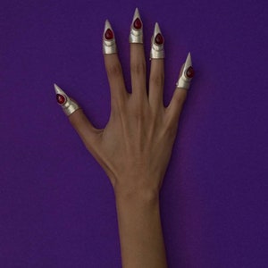 Puede incluir: Una mano con cinco anillos de plata con detalles de piedras preciosas rojas. Los anillos son puntiagudos y se asemejan a garras. La mano está sobre un fondo morado.