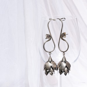 May include: A pair of silver dangle earrings with a peacock design and a flower-shaped pendant. The earrings are hanging from a glass wine glass.