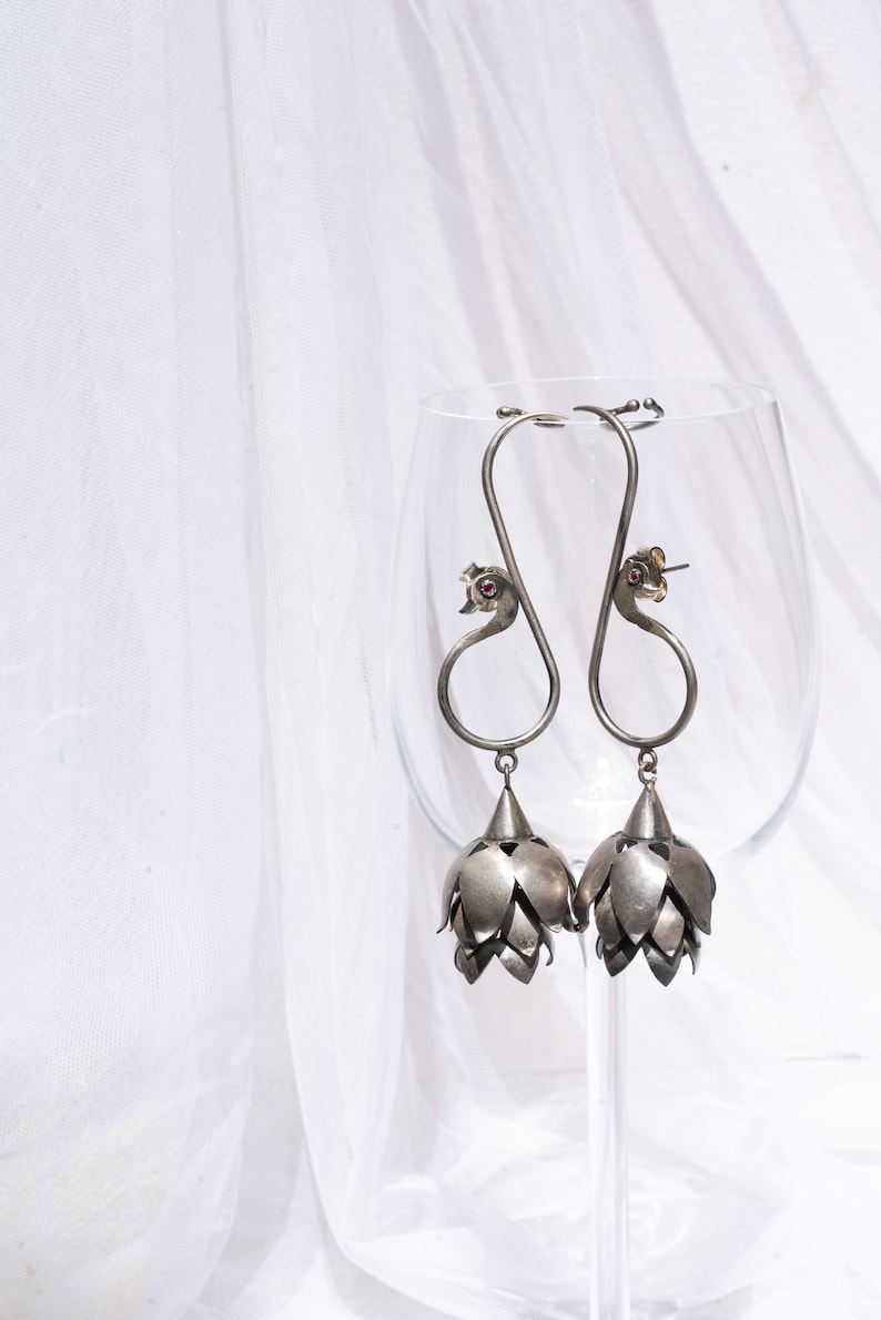 May include: A pair of silver dangle earrings with a peacock design and a flower-shaped pendant. The earrings are hanging from a glass wine glass.