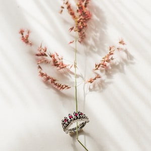 May include: A silver ring with a crown design and red gemstones. The ring is resting on a dried flower stem.