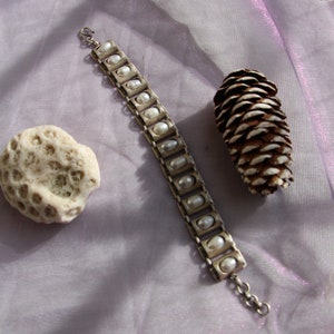 Puede incluir: Una pulsera de metal plateado que presenta una serie de eslabones rectangulares, cada uno con una peque&ntilde;a perla blanca de forma ovalada. La pulsera incluye un cierre de gancho en un extremo y una cadena de extensi&oacute;n corta.