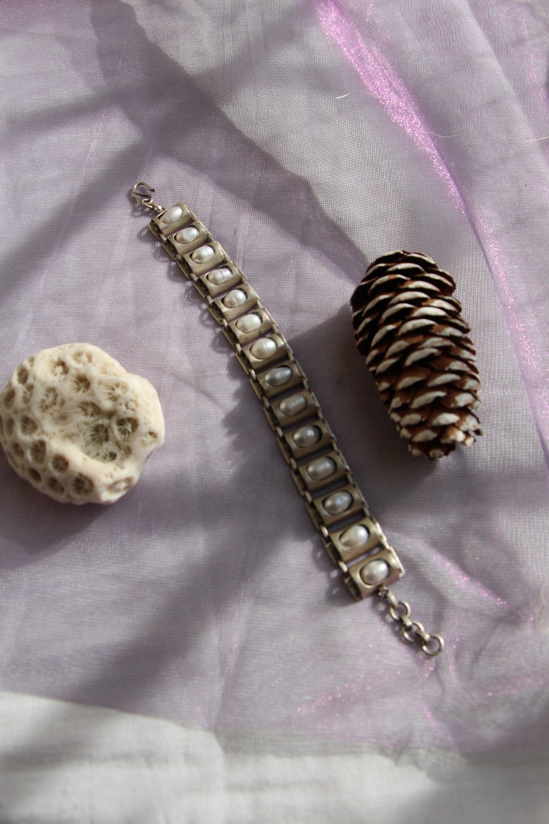 Puede incluir: Una pulsera de metal plateado que presenta una serie de eslabones rectangulares, cada uno con una peque&ntilde;a perla blanca de forma ovalada. La pulsera incluye un cierre de gancho en un extremo y una cadena de extensi&oacute;n corta.