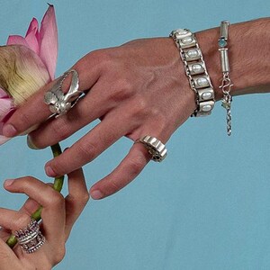 Puede incluir: Una pulsera de plata con incrustaciones ovaladas blancas, una pulsera de cadena fina de plata con una peque&ntilde;a piedra azul, un anillo de plata con dise&ntilde;o de ala, un anillo de banda ancha y un anillo de plata con piedras rosas.