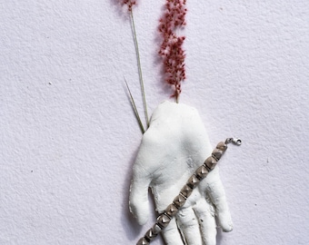 Puede incluir: Una escultura de mano de yeso blanco que sostiene dos tallos finos de hierba seca de color marrón rosáceo. Una pulsera plateada con una serie de pequeños tachones metálicos en forma de pirámide cuadrada se extiende diagonalmente sobre la palma.