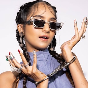 May include: Silver-framed sunglasses with dark lenses. The image features a person wearing the sunglasses, along with multiple rings and earrings. The person is also wearing a blue top. The background is plain white.