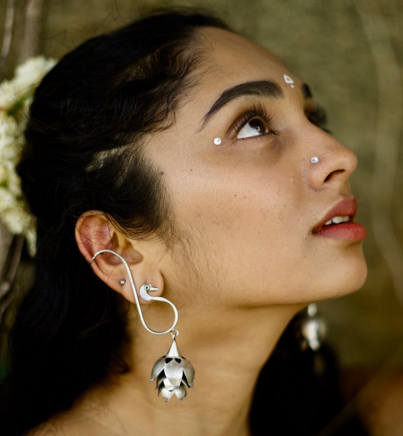 May include: A woman with dark hair and a silver nose ring wears a pair of silver earrings. The earrings are shaped like a swan with a flower-shaped pendant.