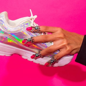 May include: A hand with pink nail polish and silver rings with turquoise stones holds a white iridescent sneaker with a white sole.