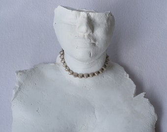 May include: A white plaster bust sculpture featuring a textured, fragmented torso and head. The sculpture is adorned with a silver-toned choker necklace composed of small, uniform, pyramid-shaped metal studs encircling the neck.