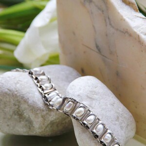 Puede incluir: Una pulsera de eslabones de metal en tono plateado que presenta una serie de marcos rectangulares, cada uno con una perla blanca de forma ovalada. La pulsera est&aacute; construida con segmentos articulados que permiten flexibilidad y un cierre seguro.