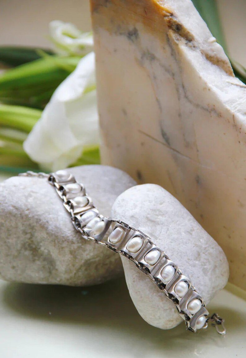 Puede incluir: Una pulsera de eslabones de metal en tono plateado que presenta una serie de marcos rectangulares, cada uno con una perla blanca de forma ovalada. La pulsera est&aacute; construida con segmentos articulados que permiten flexibilidad y un cierre seguro.