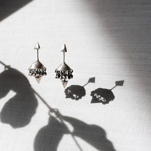 May include: A pair of silver earrings with a bell-shaped design and a pointed tip. The earrings are hanging from a white surface and casting shadows.