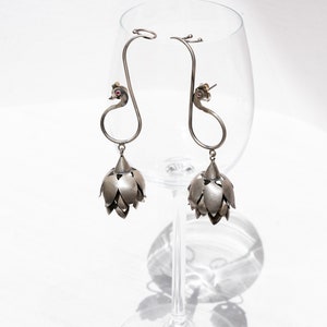 以下が含まれることがあります： 孔雀のデザインとぶら下がった花の形をしたチャームが付いた銀のイヤリングのペア。イヤリングはワイングラスからぶら下がっています。