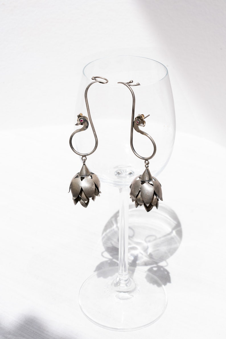 May include: A pair of silver earrings with a peacock design and a dangling flower-shaped charm. The earrings are hanging from a wine glass.