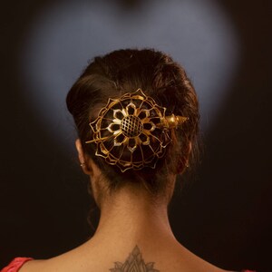 May include: A gold-toned hair accessory featuring a circular, open-work lotus flower design with a central grid pattern and a pointed pin fastener, secured in dark hair above a lotus and wing tattoo on the upper back.