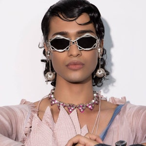 May include: A person wearing silver-framed sunglasses with black lenses, silver earrings, and a matching necklace with pink accents. The person is wearing a pink top and blue sleeves. The jewelry has a unique, geometric design.
