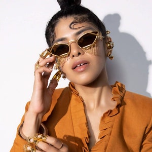 May include: Gold-coloured sunglasses with a geometric frame and dangling chain accents. The image also features ornate gold jewellery, including rings and earrings. The model is wearing an orange blazer with a ruffled collar. The overall aesthetic is bold and fashion-forward.