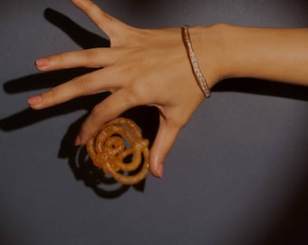 Puede incluir: Una pulsera rígida de metal en tono dorado con una serie de pequeñas piedras claras y brillantes incrustadas en la superficie exterior. La pulsera tiene una forma orgánica ligeramente ondulada y un acabado metálico reflectante.