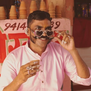 May include: A man wearing a pink shirt and heart-shaped sunglasses with rhinestones. He has a mustache and is holding a small cup in his hand. He is wearing several rings on his fingers. The background is a wall with the numbers 9414 and 59 painted on it.