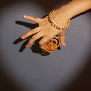 Könnte beinhalten: Eine Hand mit einem goldenen Armreif mit Diamant-ähnlichem Design hält einen Stapel goldbrauner Jalebi, ein traditionelles indisches Süßwaren.