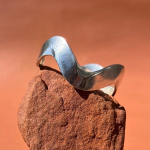 Könnte beinhalten: Ein silberner Ring mit wellenförmigem Design, der auf einem roten Stein liegt. Der Ring ist aus Metall und hat eine glatte, polierte Oberfläche.