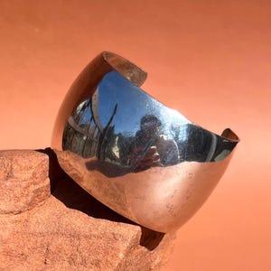 May include: A silver cuff bracelet with a smooth, rounded surface. The bracelet is reflecting the surrounding environment.