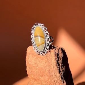 Op de afbeelding: Een zilveren ring met een ovale steen gezet in een filigraan ontwerp. De steen is een mix van bruine, gele en witte kleuren.